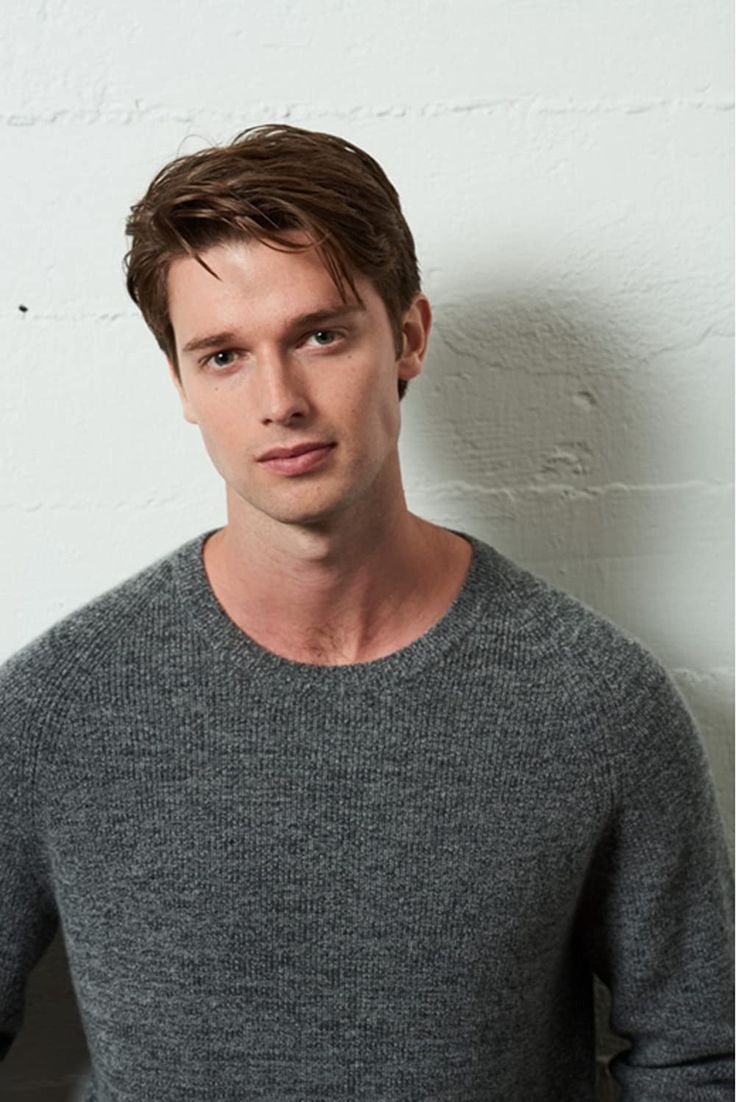 a man standing in front of a white wall wearing a gray sweater and black pants