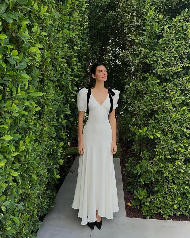 a woman wearing a white dress standing in front of some bushes