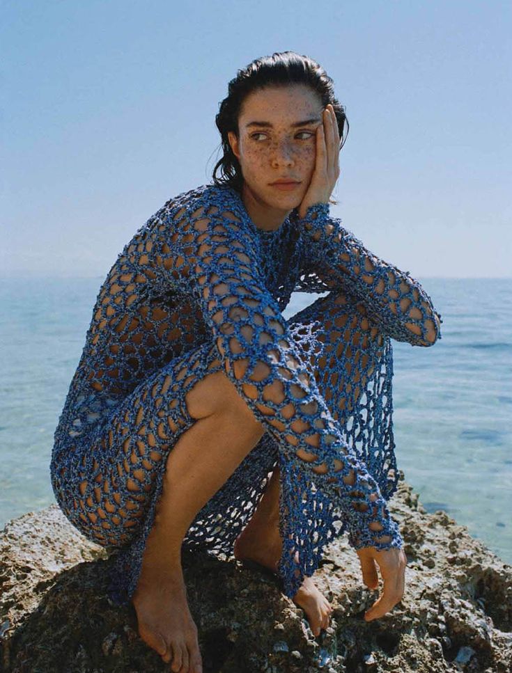 a woman sitting on top of a rock next to the ocean holding her hand near her face