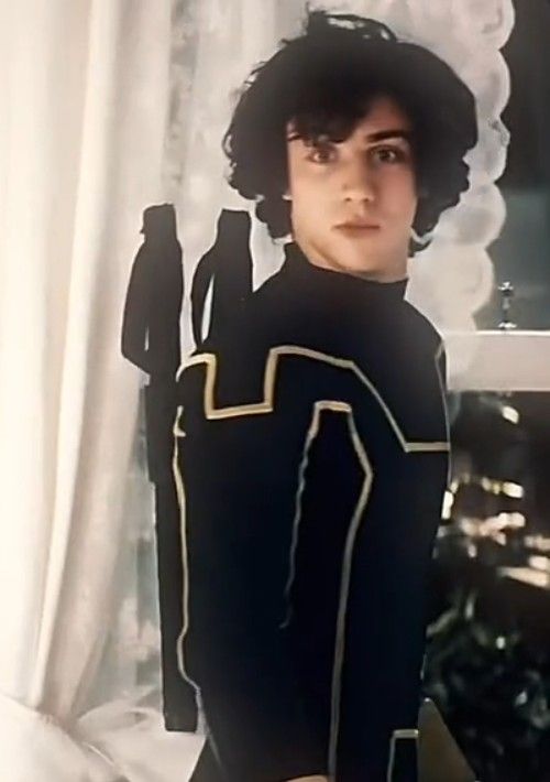 a young man dressed in black and gold standing next to a window with white curtains