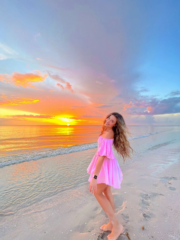 a beautiful young woman standing on top of a beach next to the ocean at sunset