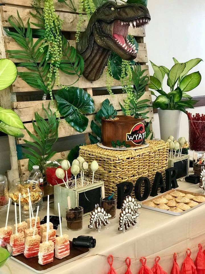 a table topped with lots of desserts and plants next to a wall mounted dinosaur head