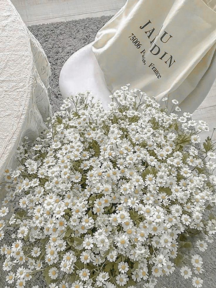 white daisies in a vase on the floor next to a chair and bag with an embroidered name tag