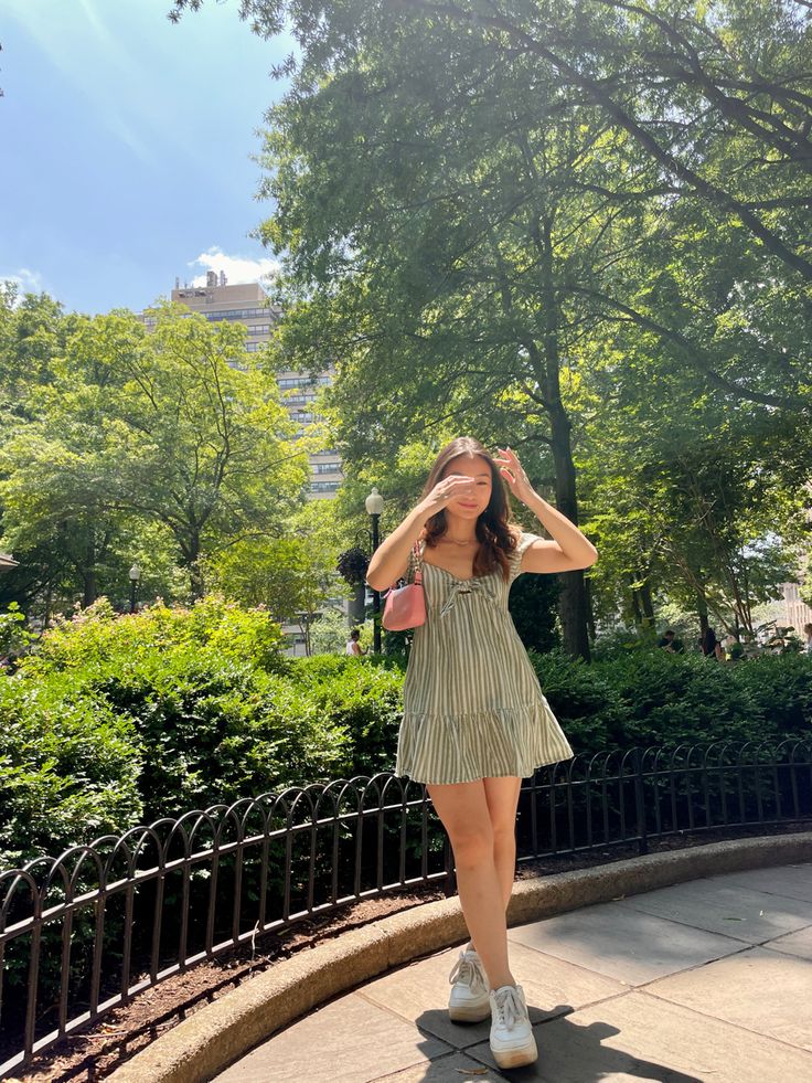 a woman in a dress is standing on the sidewalk and looking through her sunglasses with trees behind her