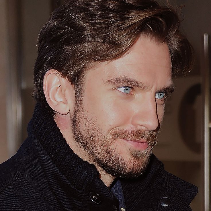 a close up of a person wearing a coat and tie with blue eyes looking at the camera