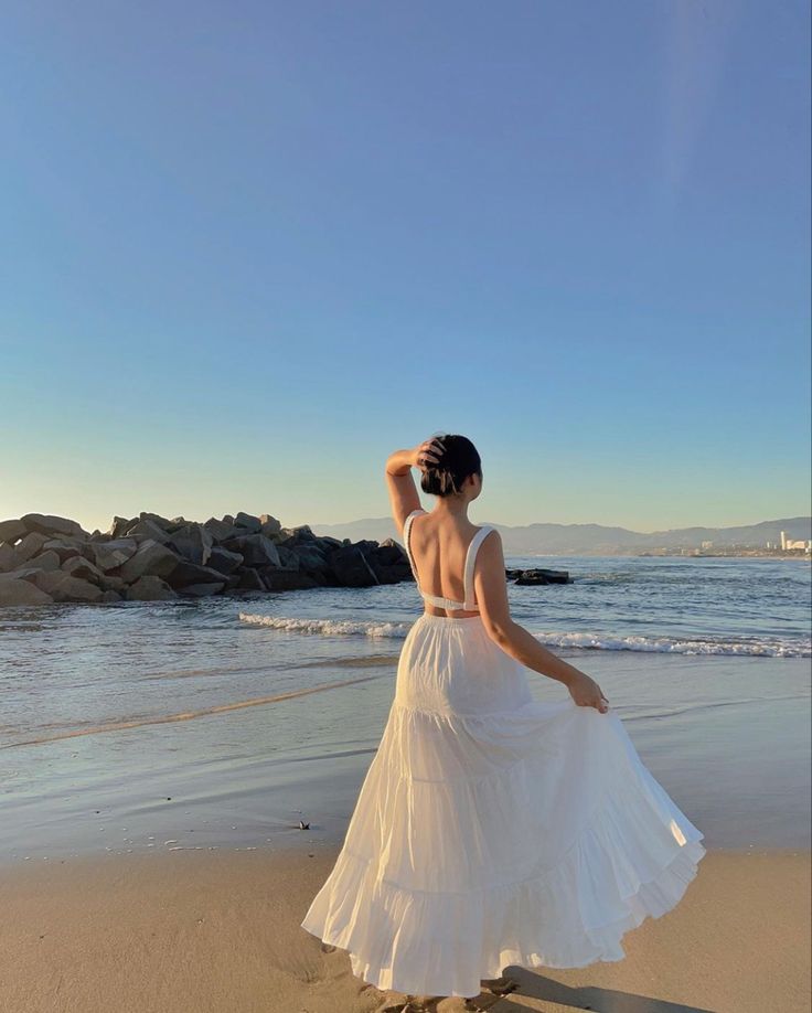 a woman standing on top of a sandy beach next to the ocean wearing a white dress