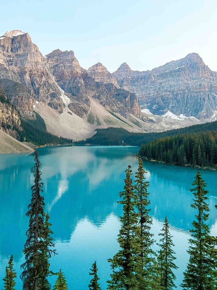 a lake surrounded by mountains and trees with blue water in the foreground on a sunny day