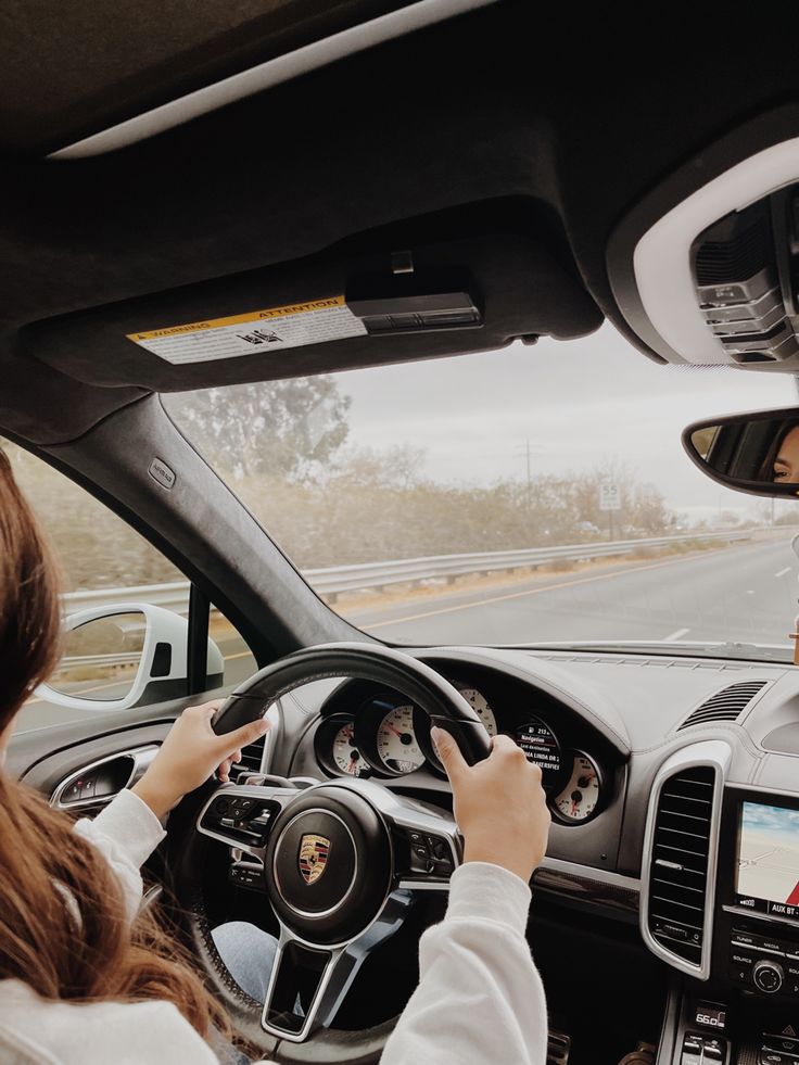 a woman driving a car on the road with her hand on the steering wheel and another person sitting in the driver's seat