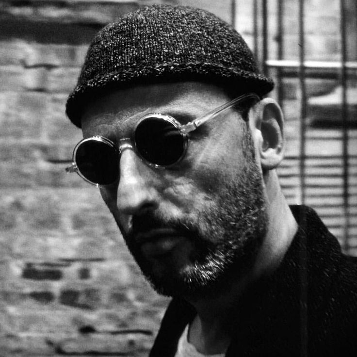 a man with sunglasses and a hat on standing in front of a brick wall looking at the camera
