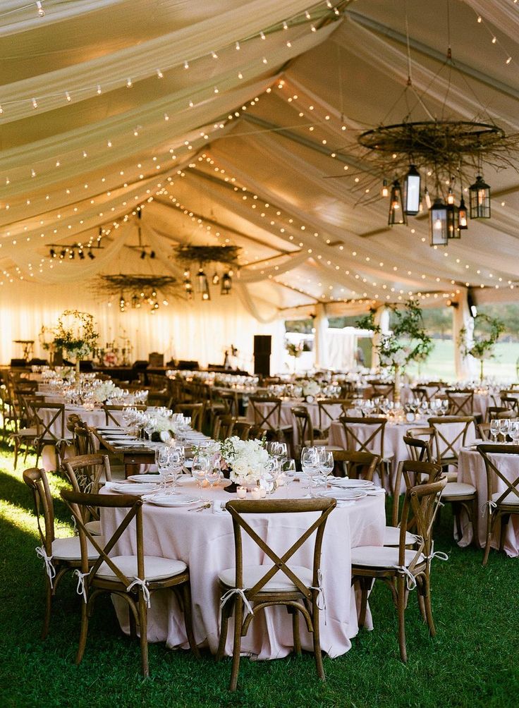 tables and chairs are set up under a tent for an outdoor wedding reception with lights strung from the ceiling