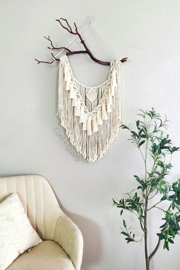 a white wall hanging next to a chair and tree branch in a living room with a potted plant