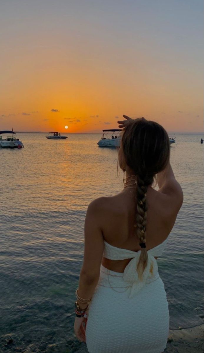 a woman in a white dress looking out at the ocean with boats on the water