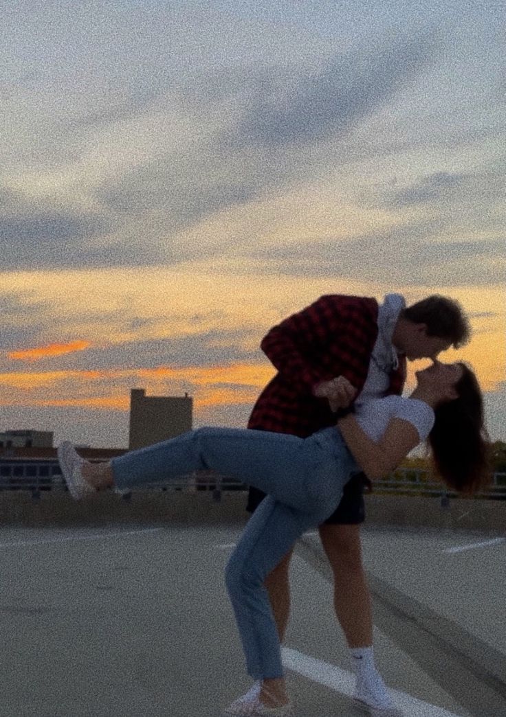 a man and woman kissing while standing on top of a parking lot with the sun setting behind them