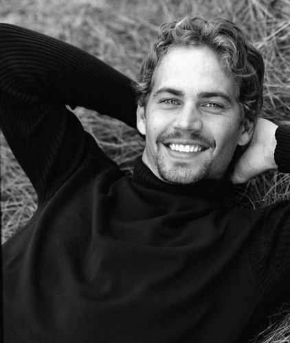 a black and white photo of a smiling man with his hands on his head, wearing a turtle neck sweater