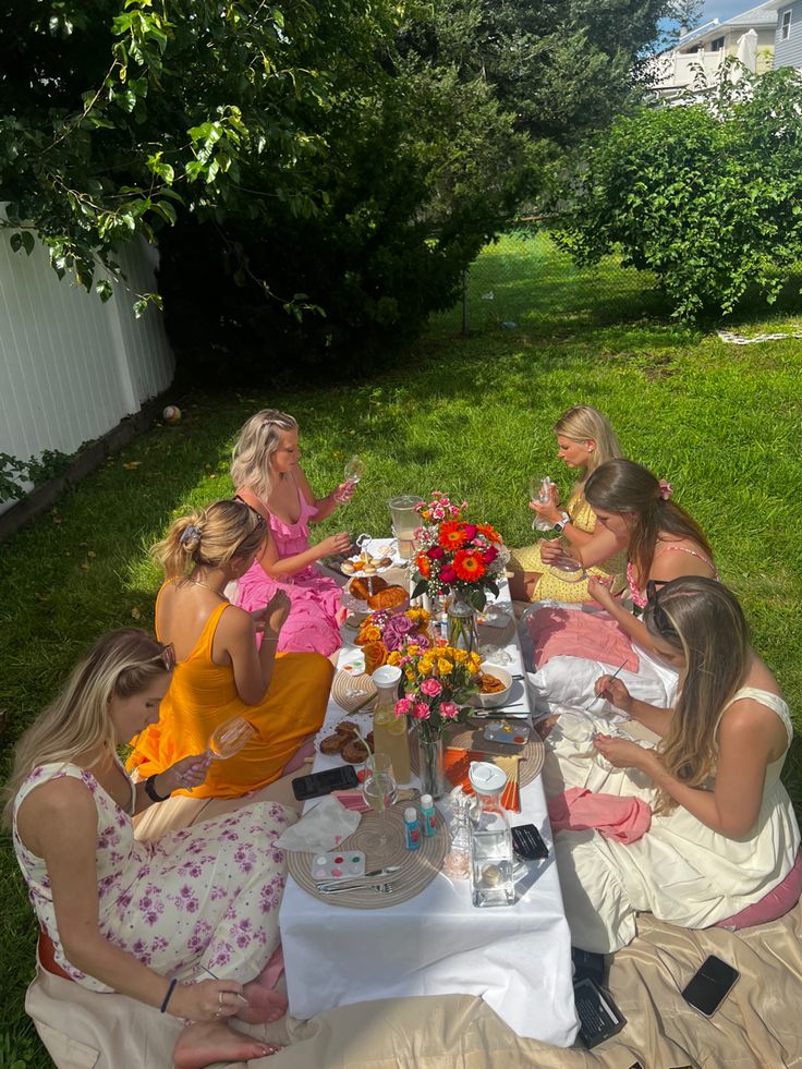 a group of women sitting around a table with food and drinks on it in the grass