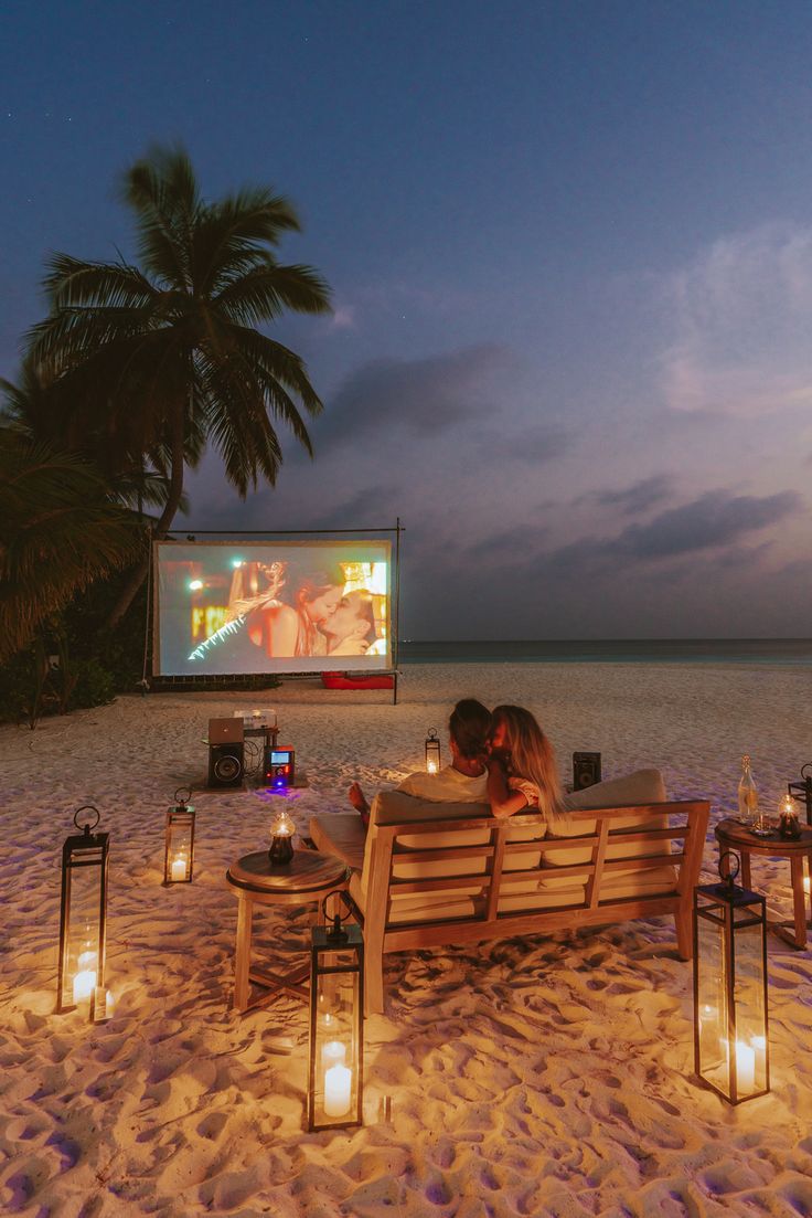 two people sitting on a bench in the sand watching a movie with candles lit up