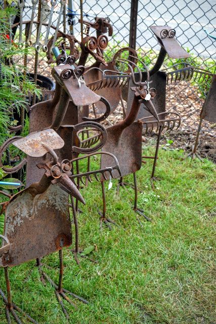 several rusty metal sculptures sitting in the grass near a chain link fence and some bushes