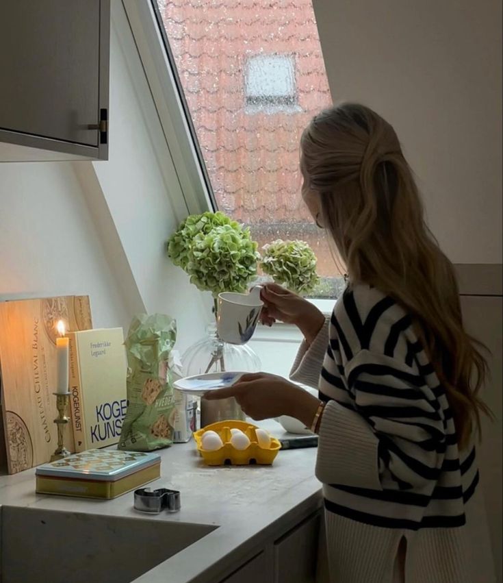 a woman standing in a kitchen next to a window with flowers on the windowsill