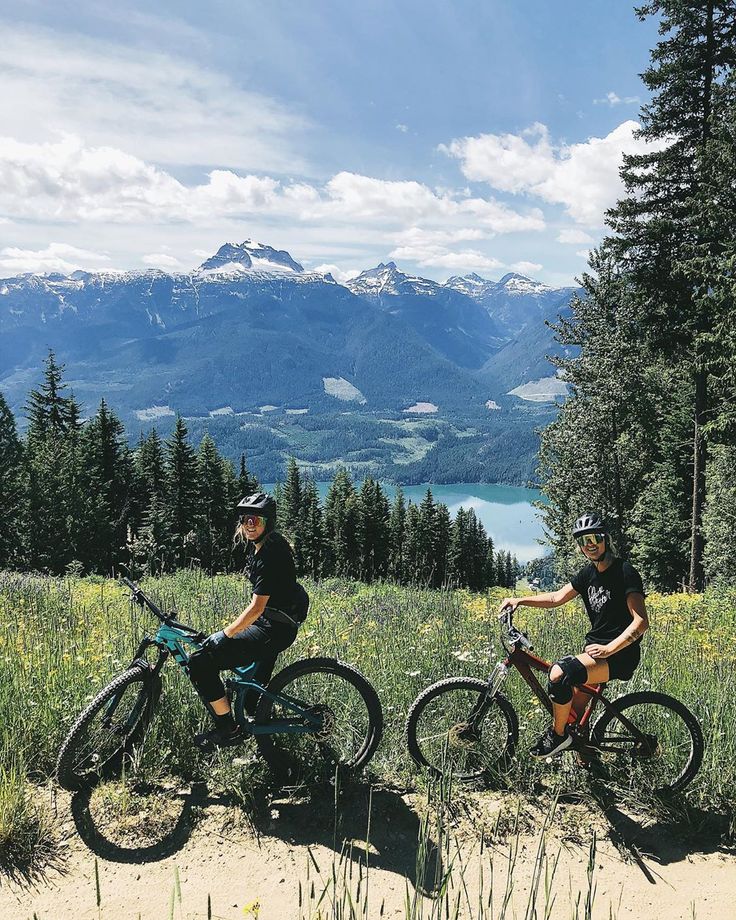 two people riding bikes on top of a dirt hill next to trees and grass with mountains in the background