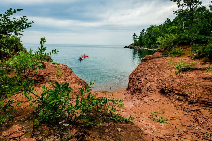 a person on a kayak in the water near some rocks and trees, with another boat