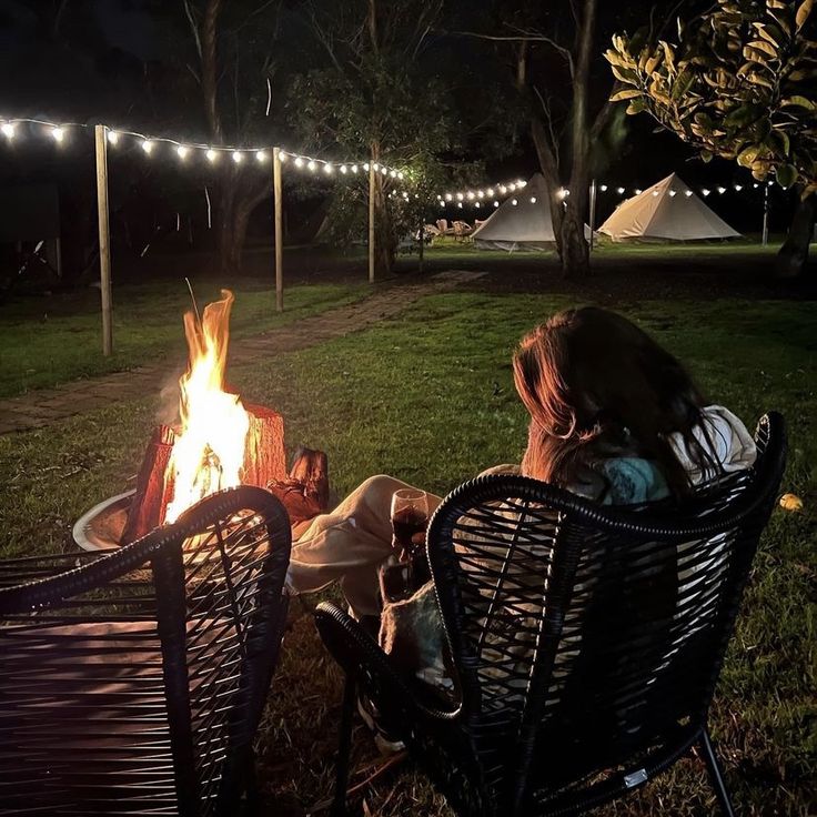 two people sitting in chairs around a fire pit at night with lights strung across the lawn