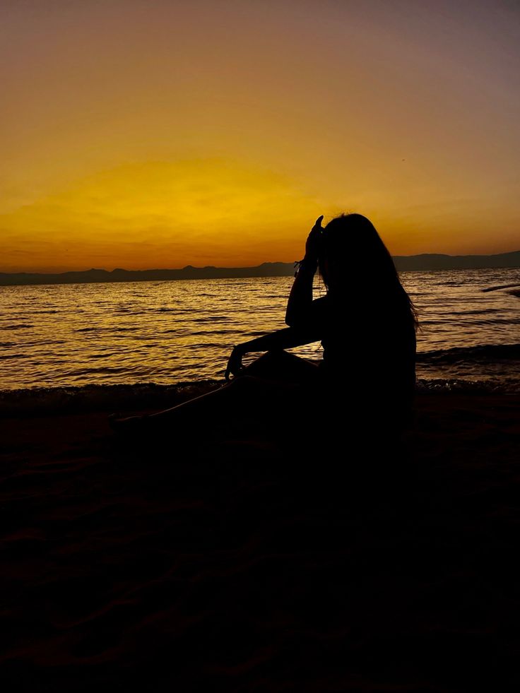 a woman sitting on top of a sandy beach next to the ocean at sunset or dawn