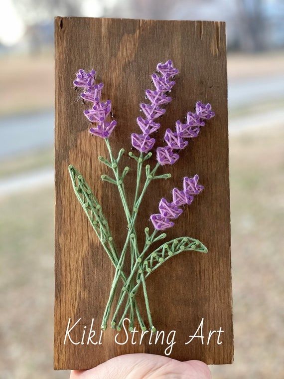 a hand holding up a piece of wood with purple flowers on it and the words, kiki string art