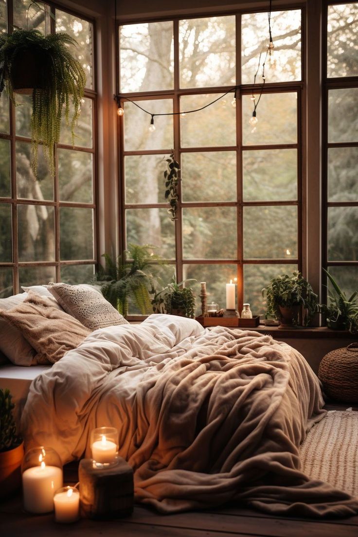 an unmade bed with candles on the floor in front of large windows and potted plants