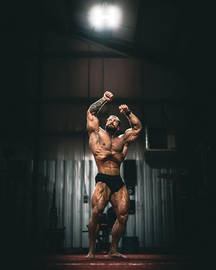 a bodybuilding man flexing his muscles in the dark, with one arm raised