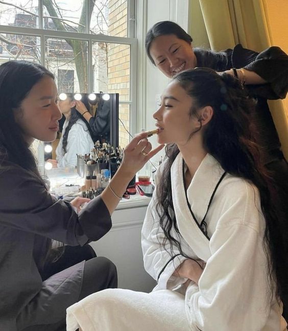 two women are getting their makeup done in front of a window while another woman looks on