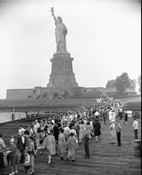 an old photo of people walking on a pier in front of the statue of liberty