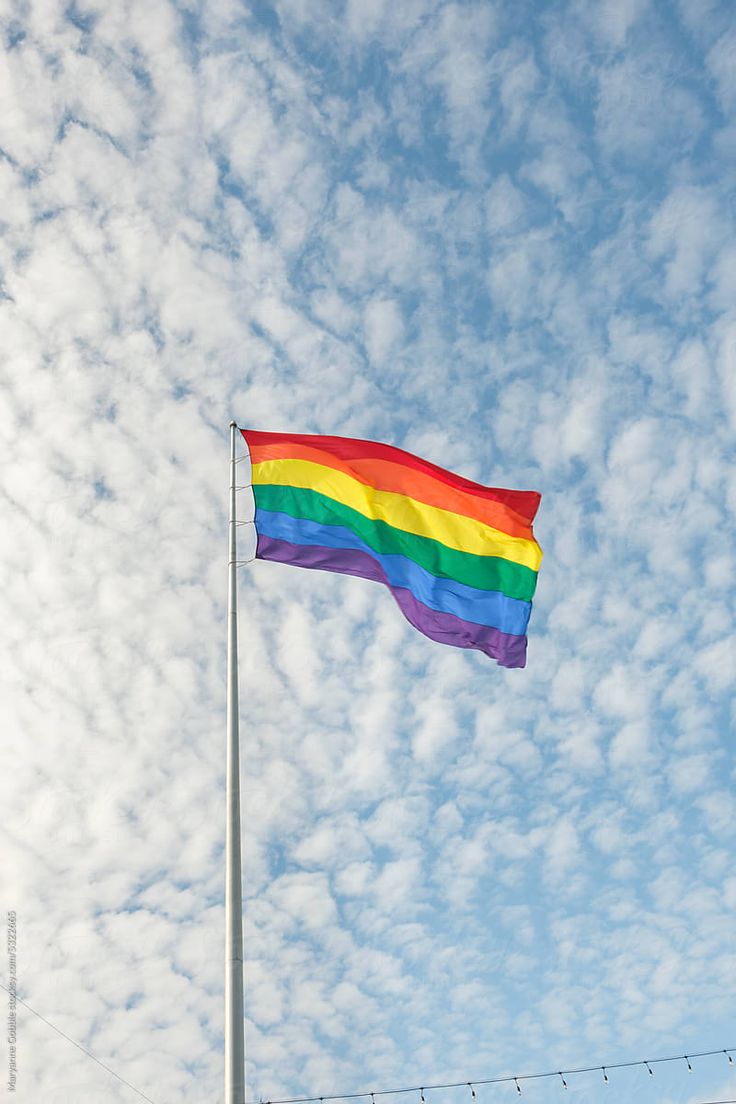 a rainbow flag is flying high in the blue sky with some white clouds behind it