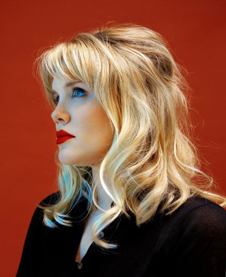 a woman with long blonde hair and blue eyes looks off into the distance while standing against a red background