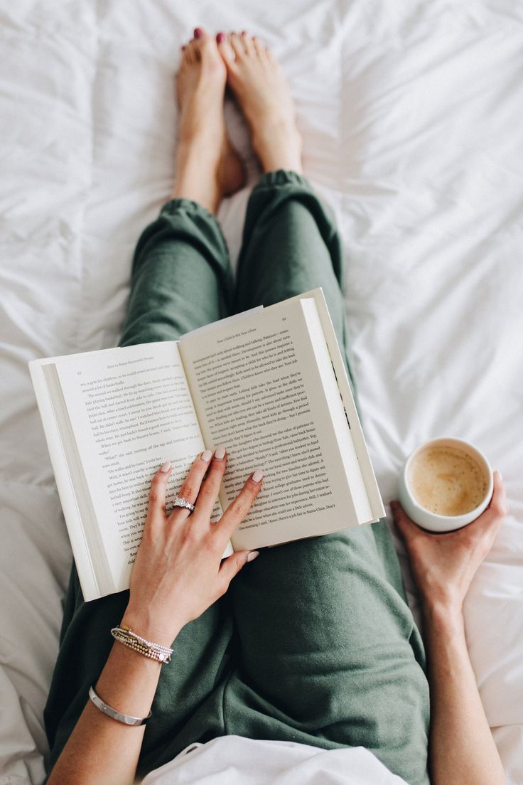 a person laying in bed reading a book and holding a cup of coffee with both hands