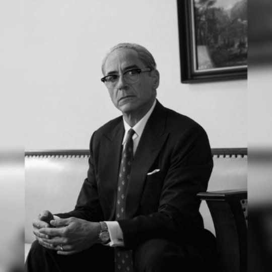 a man in a suit and tie sitting on a couch with his hands folded out