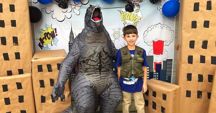 two boys standing in front of a cardboard godzilla costume with balloons on the wall behind them