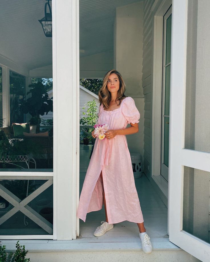 a woman in a pink dress is standing on the front porch with flowers and smiling