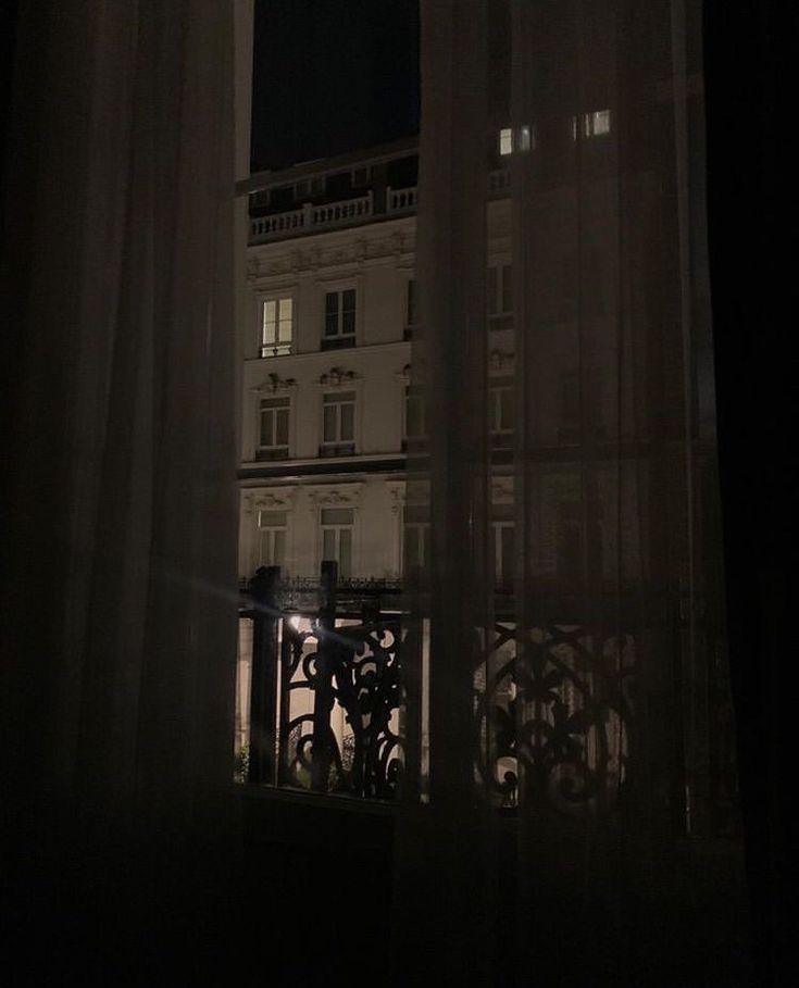 an open window at night with the view of a building through it's sheer curtains