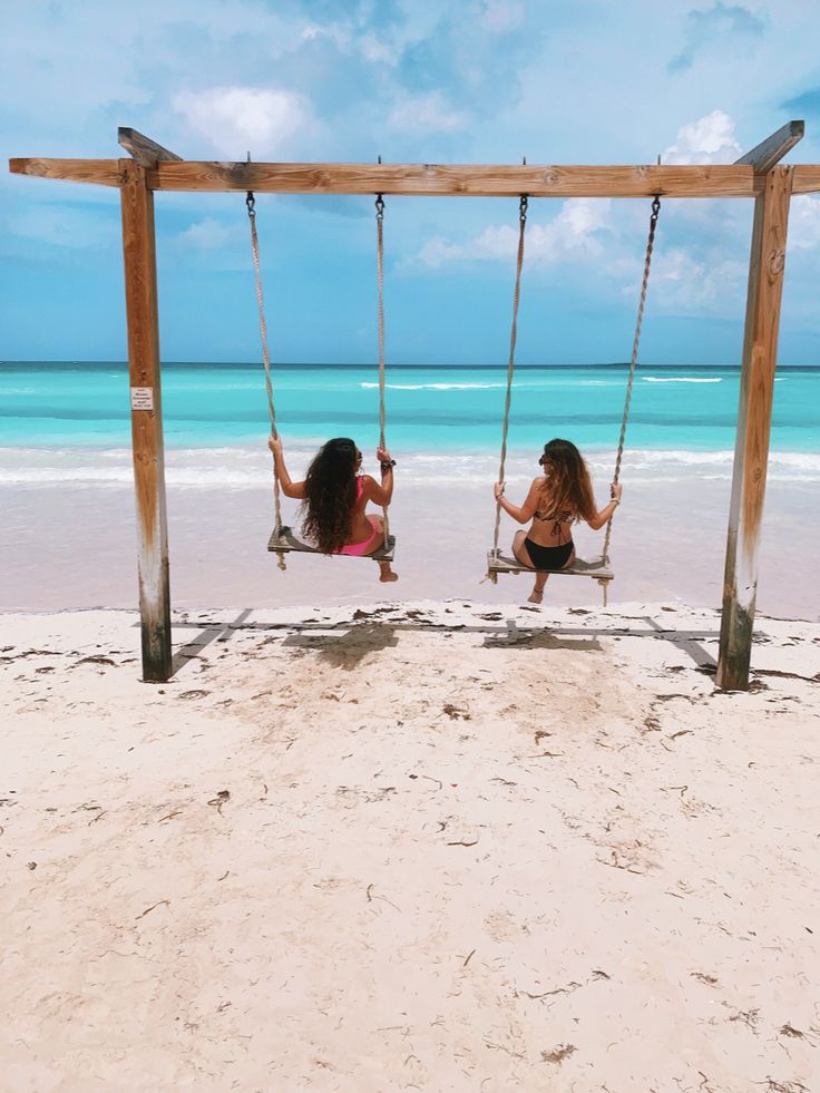 two women sitting on swings in the sand at the beach, one holding onto another