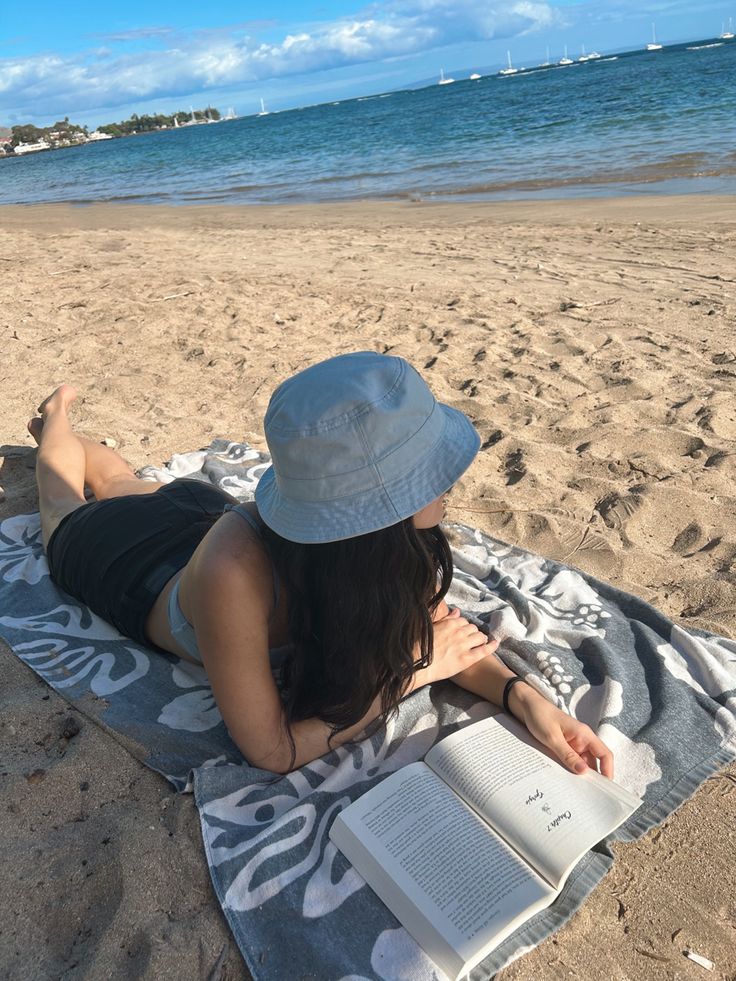 a woman laying on top of a beach next to the ocean with an open book