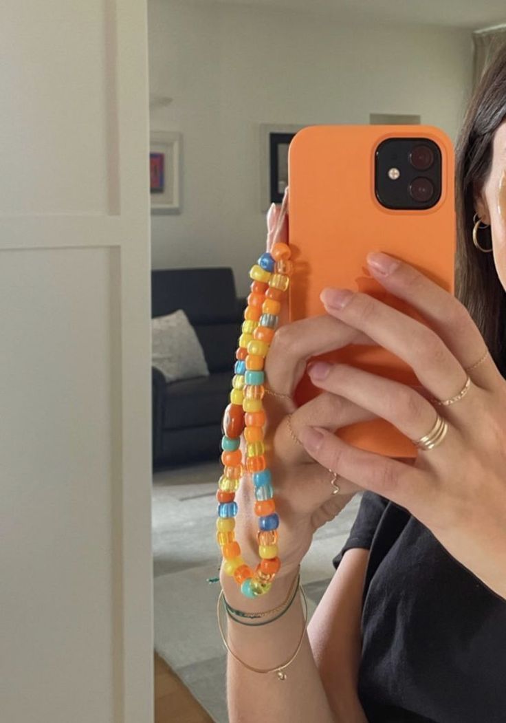 a woman taking a selfie in her living room with an orange phone case and beaded bracelets