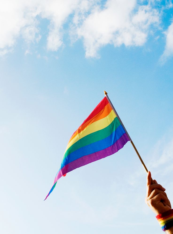 a person holding a rainbow flag in their hand under a blue sky with white clouds