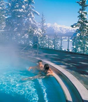 two people in a hot tub surrounded by snow covered trees and water with steam rising from it