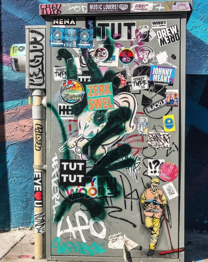 a parking meter covered in lots of stickers next to a wall with graffiti on it