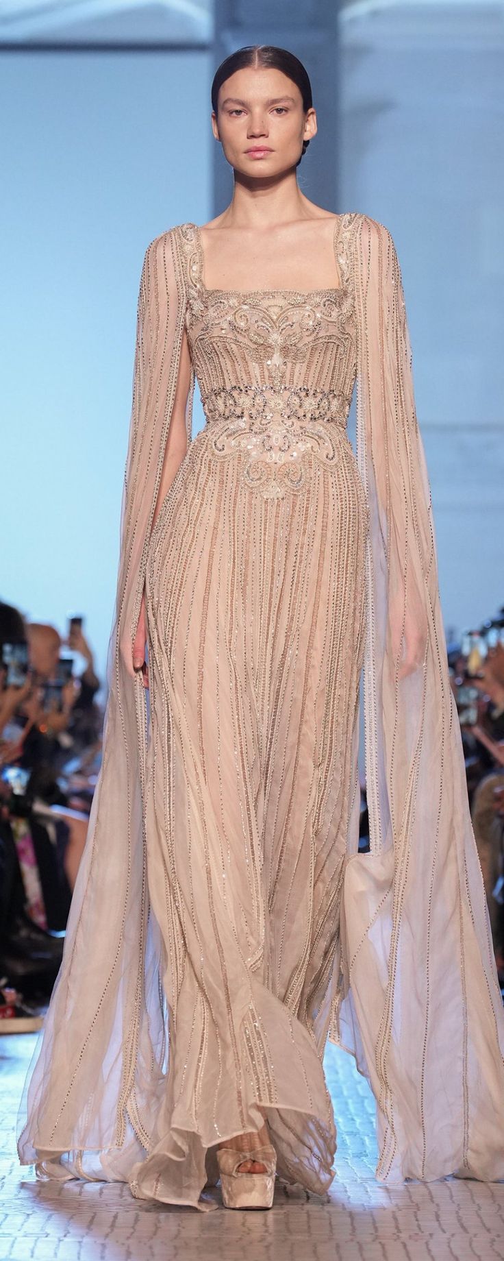 a model walks down the runway in a beige gown with sheer sleeves and an embellished cape