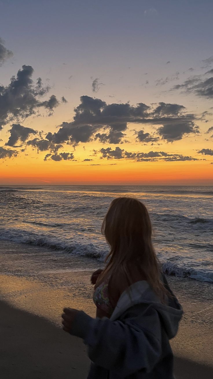 a woman standing on top of a sandy beach next to the ocean at sunset with clouds in the sky