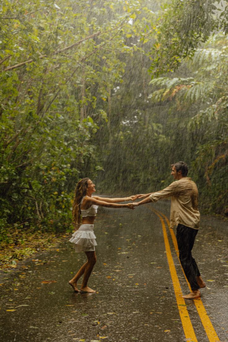 two people holding hands in the rain on a road with trees and bushes behind them