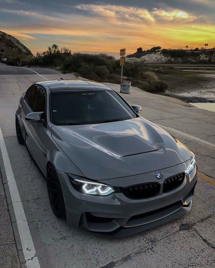 a grey bmw is parked on the side of the road near water and hills at sunset