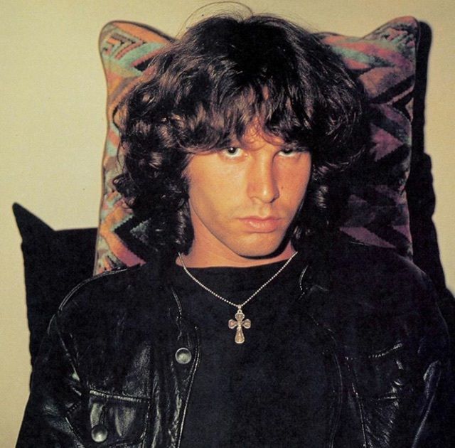 a man with long hair wearing a black leather jacket sitting in front of a pillow