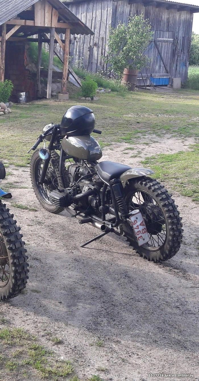 two motorcycles are parked in the dirt near a barn and shed, one has a helmet on it's back end